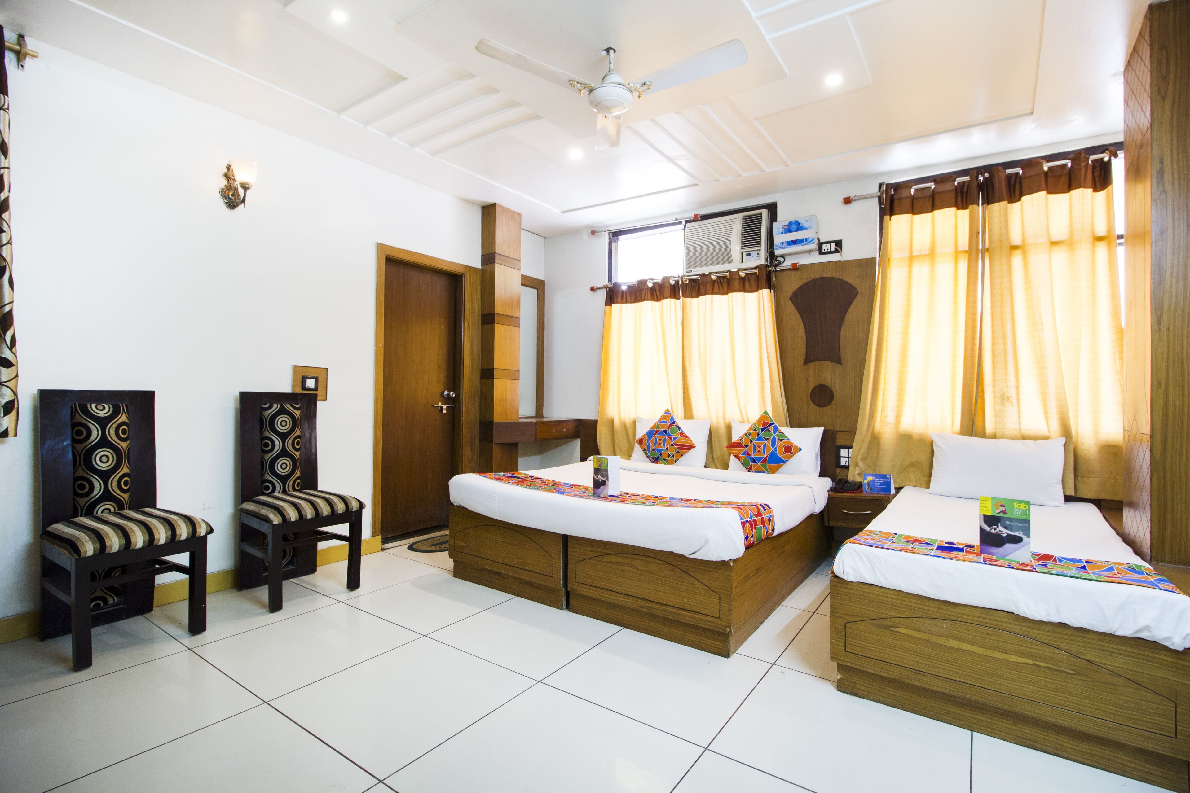 A hotel room with two beds, a ceiling fan, an air conditioning unit, and a window with curtains.
