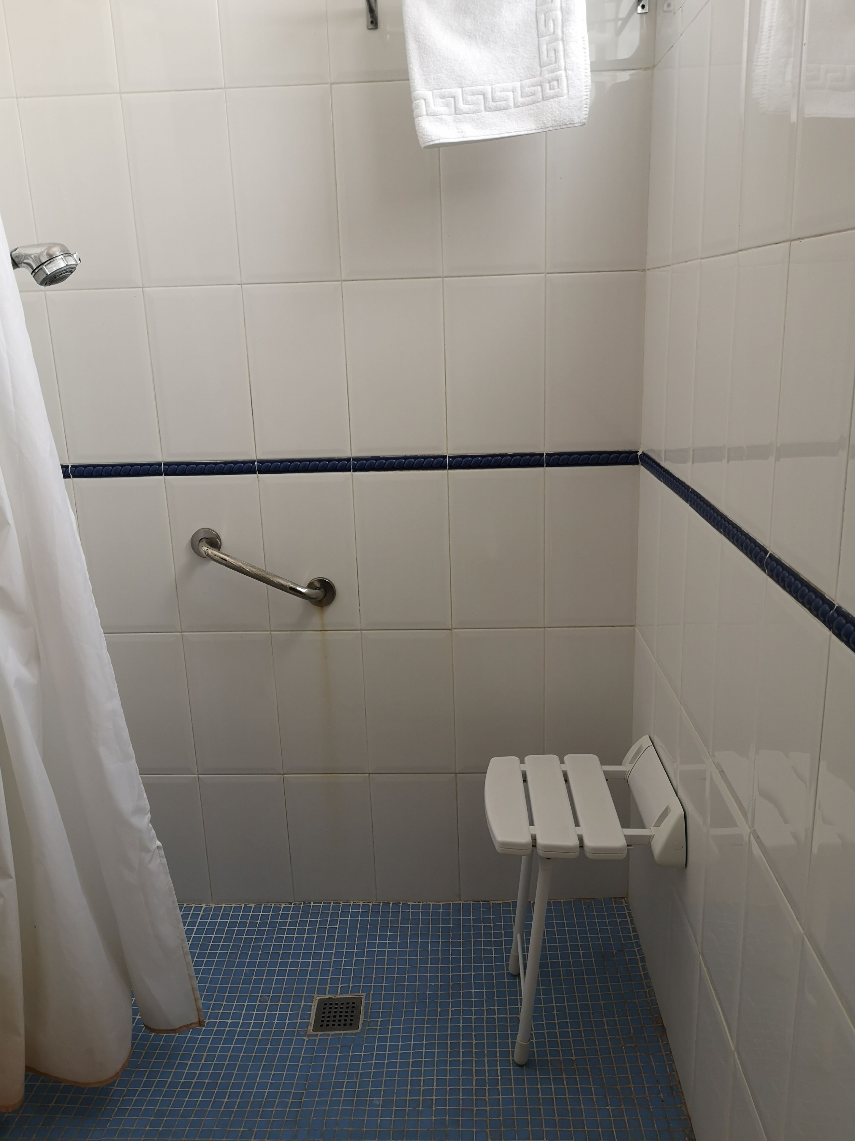 Une petite salle de bain carrelée, équipée d’une douche, d’une barre d’appui et d’un banc blanc.
