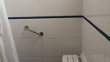 Une petite salle de bain carrelée, équipée d’une douche, d’une barre d’appui et d’un banc blanc.