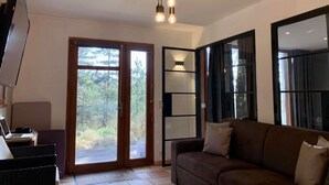 A living room with a brown sofa, a TV on a stand, and a laptop on a table. There is a sliding glass door leading to a balcony with a view of trees. - La Villa dans les Pins (Quend)