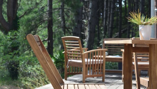 A wooden deck with a table and chairs, surrounded by a forested area. - La Villa dans les Pins (Quend)