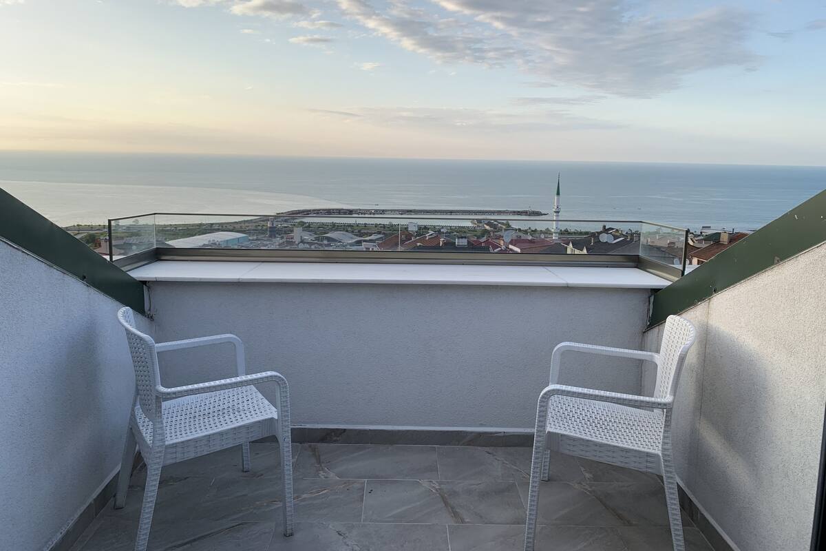 A balcony with two white wicker chairs, a view of the sea, and a distant cityscape.