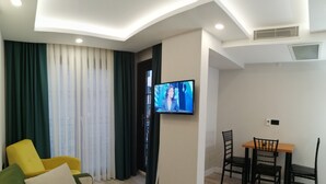 A modern living room with a flat-screen TV mounted on the wall, a green sofa, a yellow armchair, and a dining area with a wooden table and black chairs. - White Golden Suite Hotel (Trabzon)