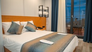 A hotel room with a wooden headboard, a bed with patterned bedding, a bedside table with a phone, and a view of the outdoors through a glass door. - White Golden Suite Hotel (Trabzon)