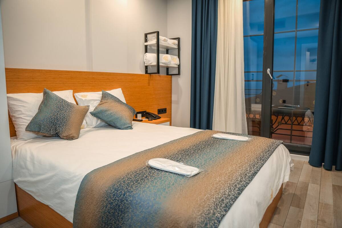 A hotel room with a wooden headboard, a bed with patterned bedding, a bedside table with a phone, and a view of the outdoors through a glass door.