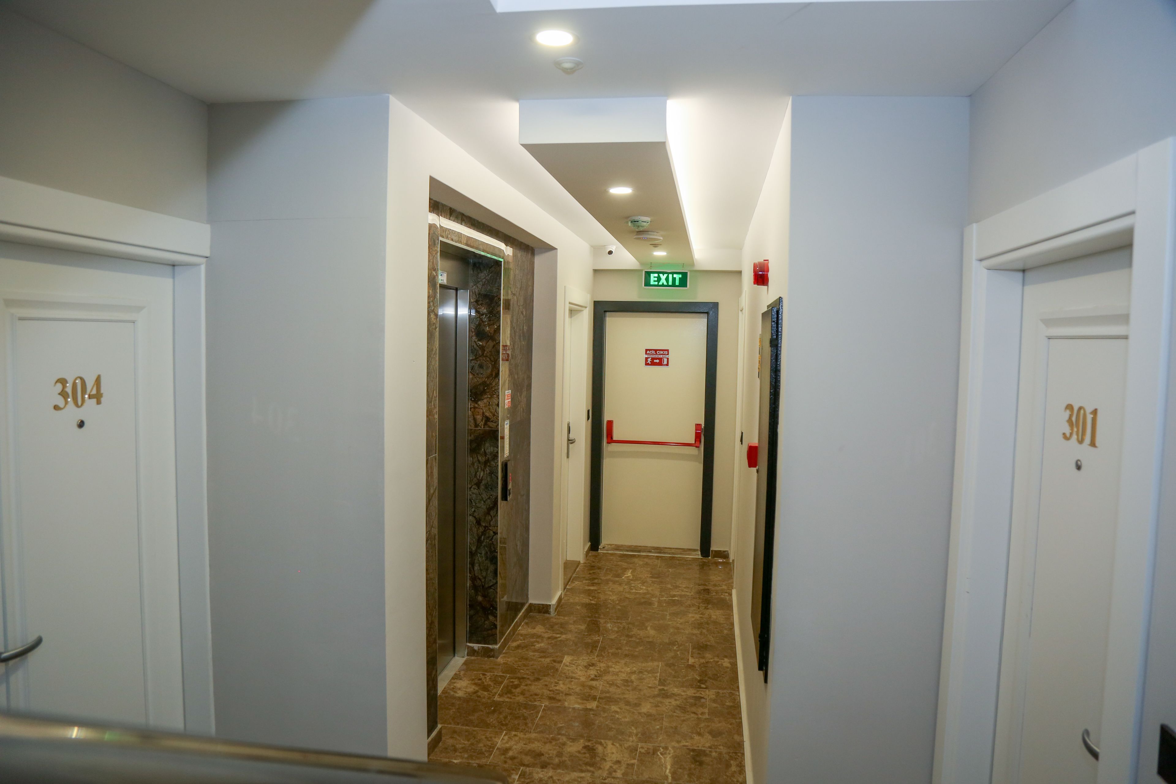 Un couloir d’hôtel avec les portes numérotées 304 et 301, un ascenseur et un panneau indiquant la sortie.