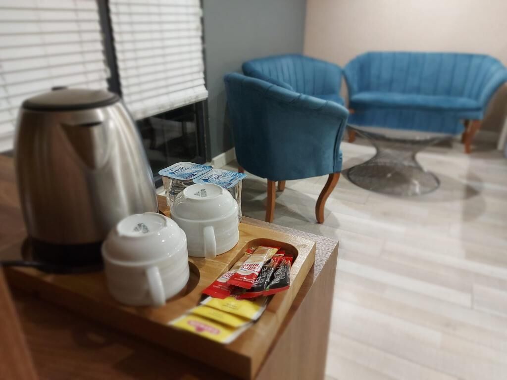 A breakfast tray with a coffee maker, cups, and snacks on a wooden table in a room with blue chairs and a window with blinds.