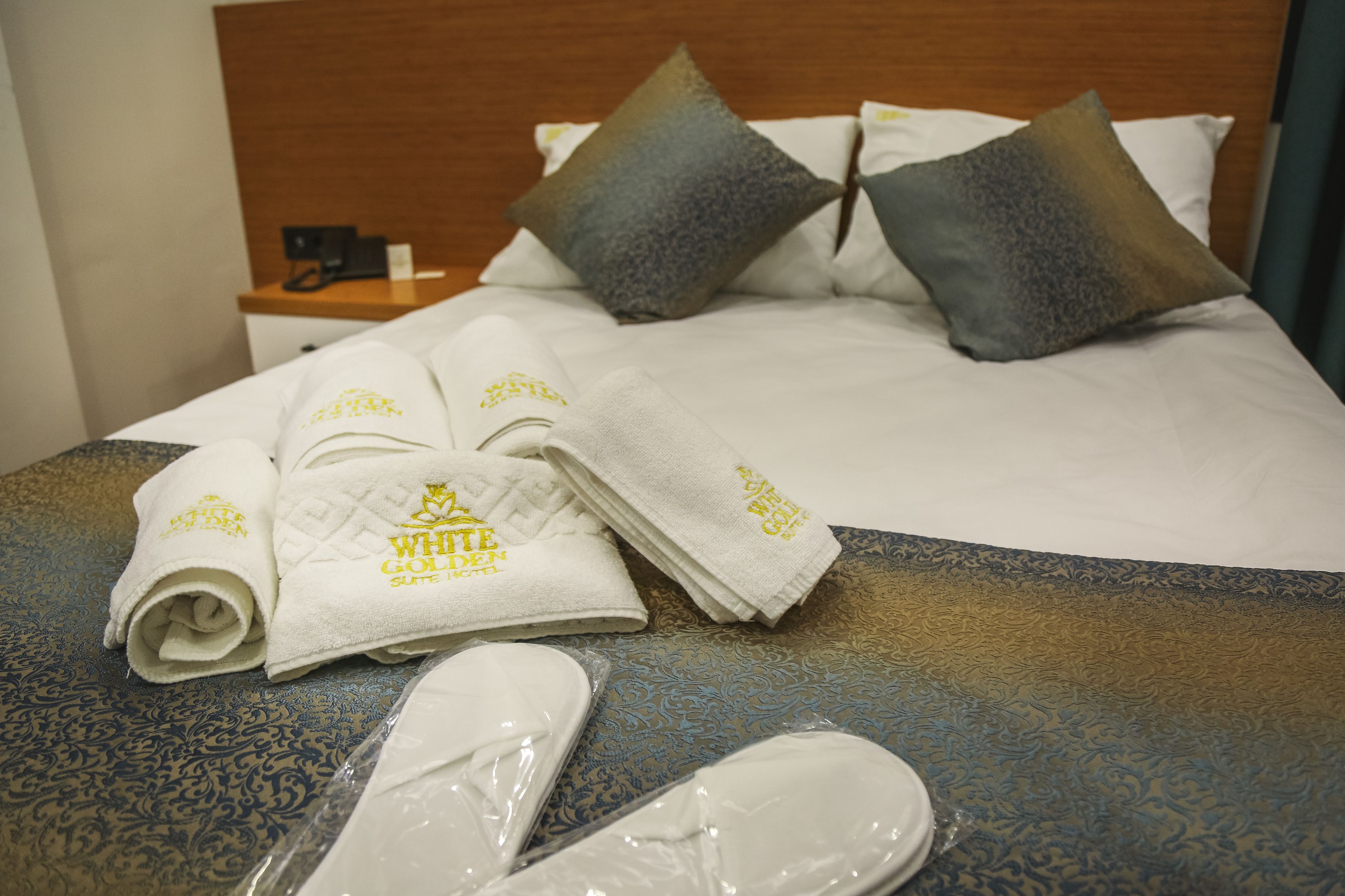 A neatly made bed with white linens and two pillows, a wooden headboard, and a bedside table with a phone and a lamp.
