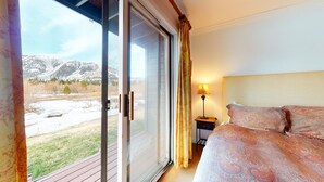 Ein Schlafzimmer mit einem Bett, einem Nachttisch, einer Lampe und einer Glasschiebetür, die zu einer verschneiten Außenanlage mit Bergen im Hintergrund führt.