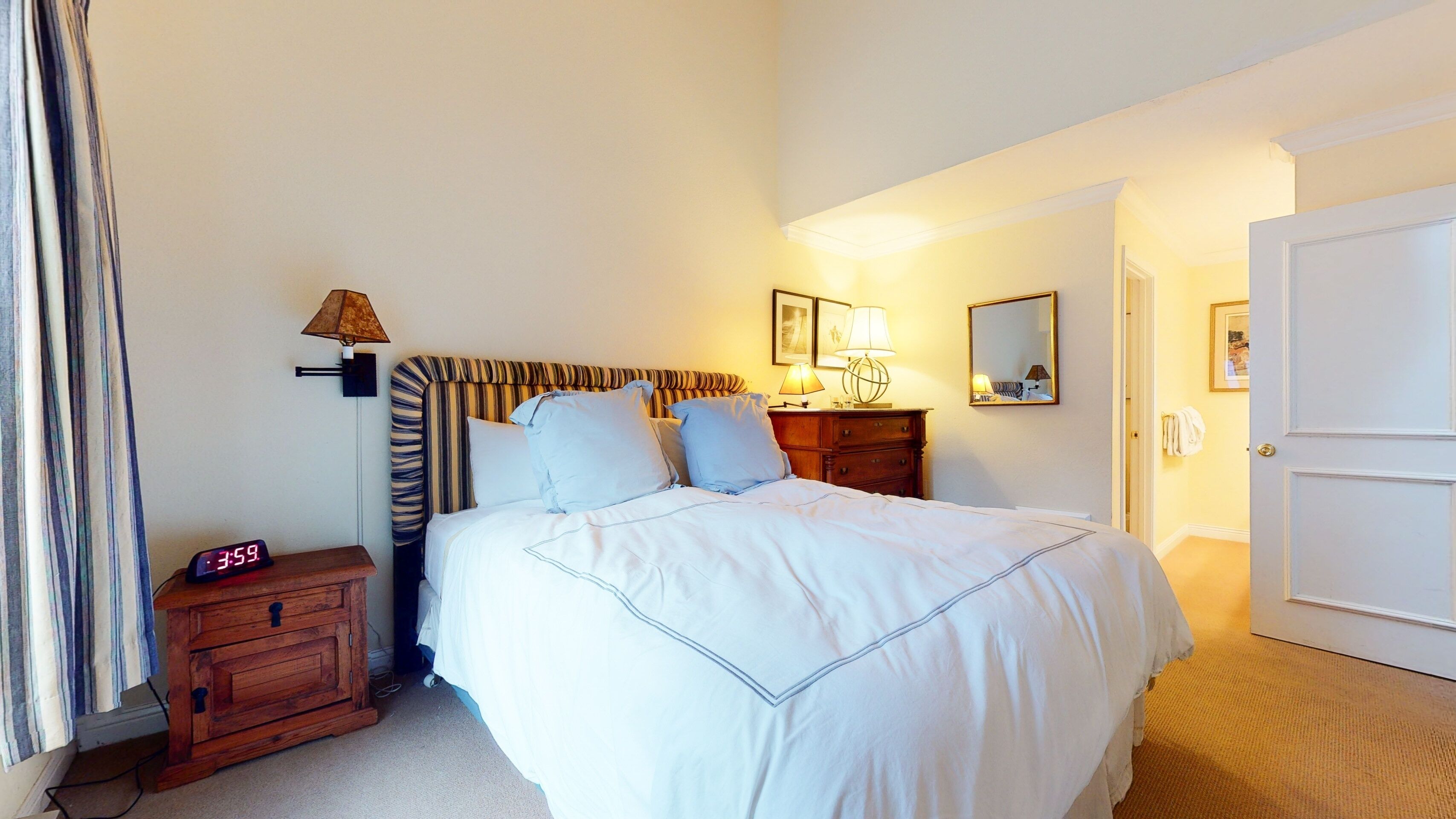 Une chambre d’hôtel avec un grand lit, une table de chevet avec une lampe, une commode en bois, un miroir et une porte.