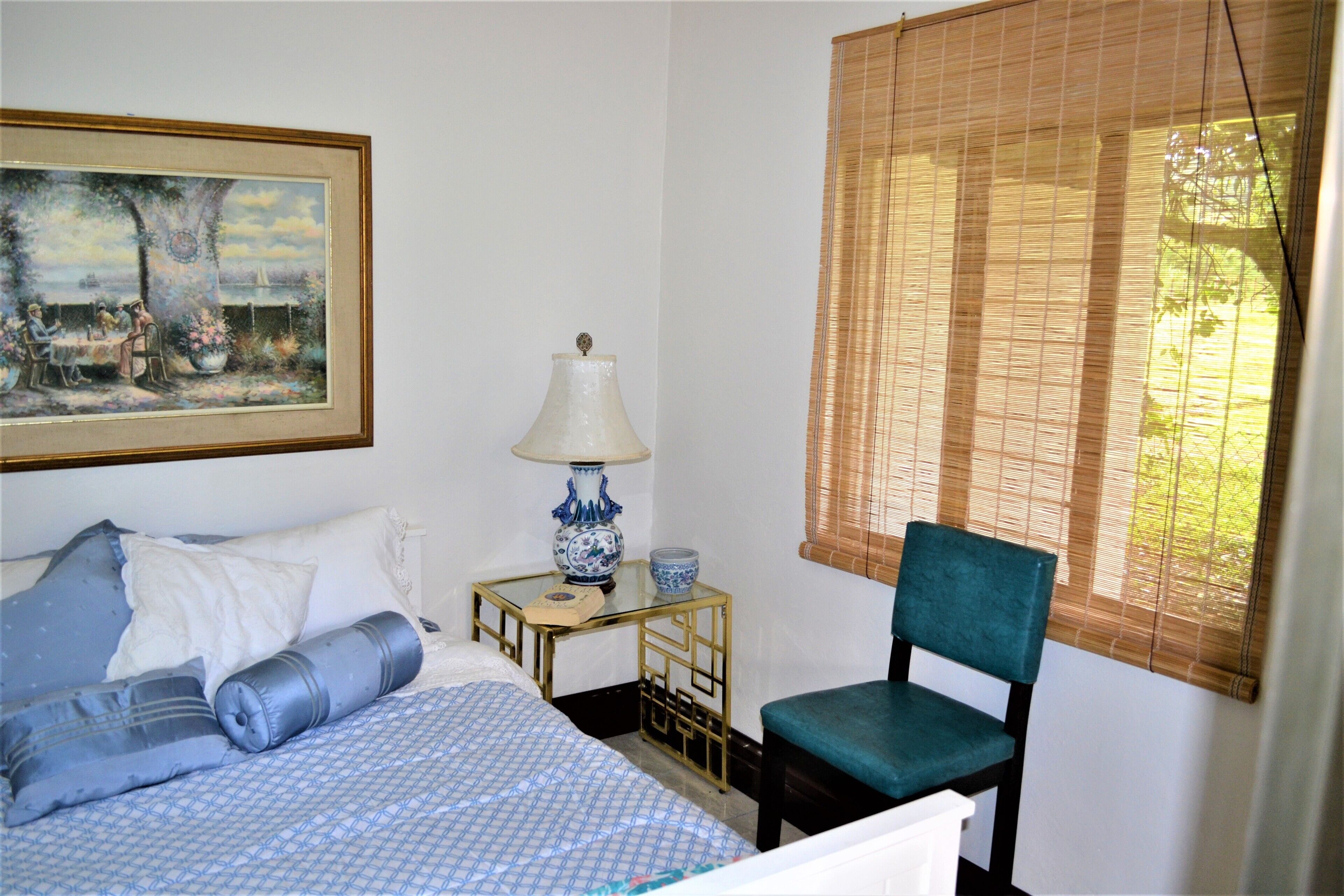Un dormitorio con cama, una silla, una mesa con lámpara y un cuadro en la pared.