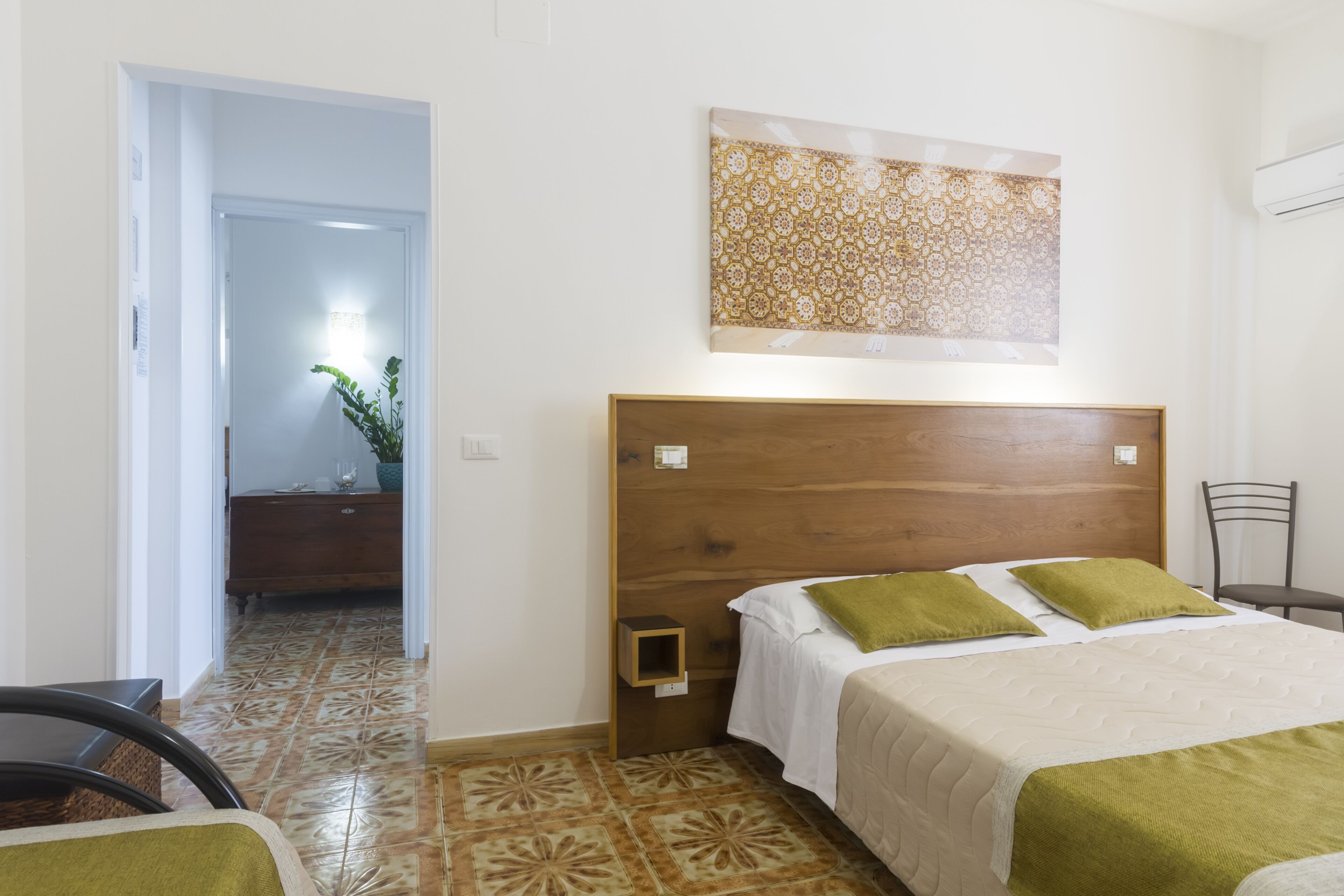 Habitación de hotel con cabecera de madera, cama con almohadas verdes, mesita de noche y una obra de arte colgada en la pared.