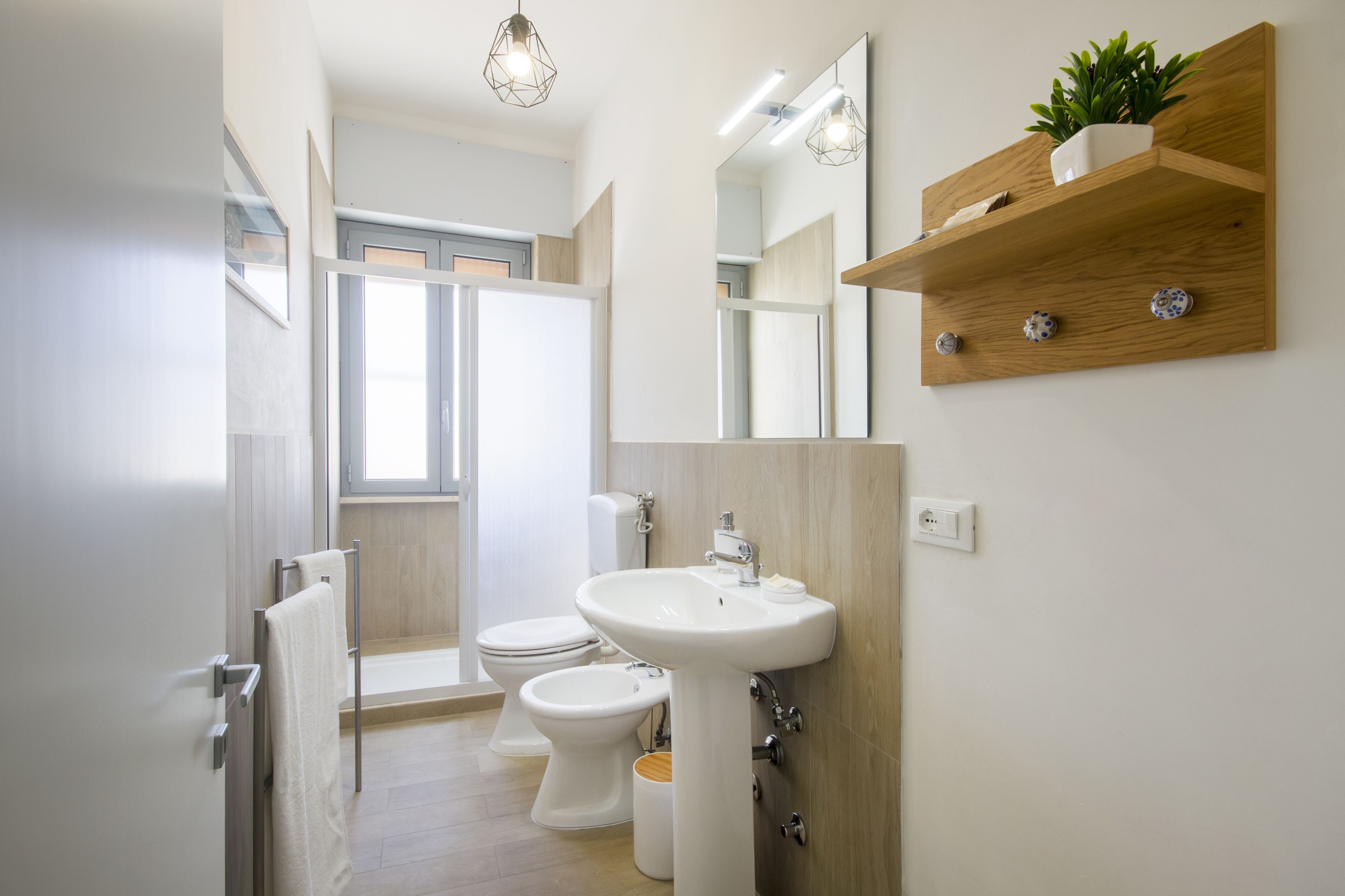 Een moderne badkamer met toilet, bidet en wastafel. Er is een houten plankje met een plant en een spiegel boven de wastafel.