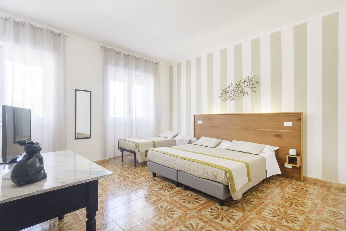 Quarto de hotel com cama, escrivaninha, cadeira, televisão e um objeto decorativo na parede.