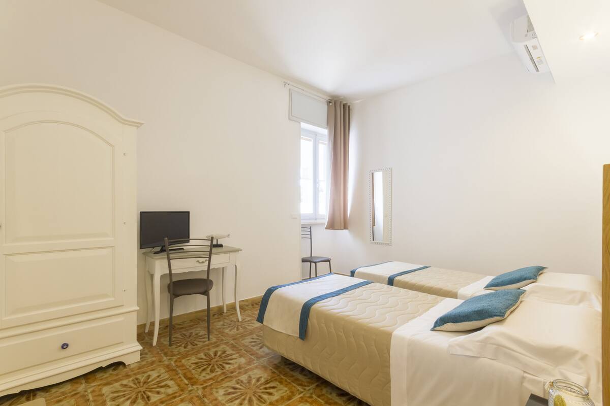 Quarto de hotel com duas camas, uma escrivaninha com computador, um guarda-roupa e uma janela com cortinas.