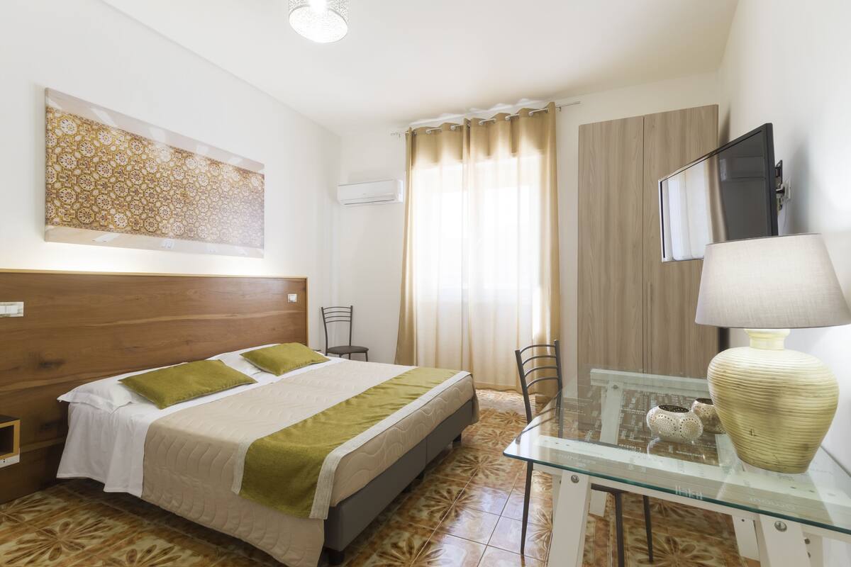 Quarto de hotel com cama, escrivaninha com luminária, televisão e janela com cortinas.
