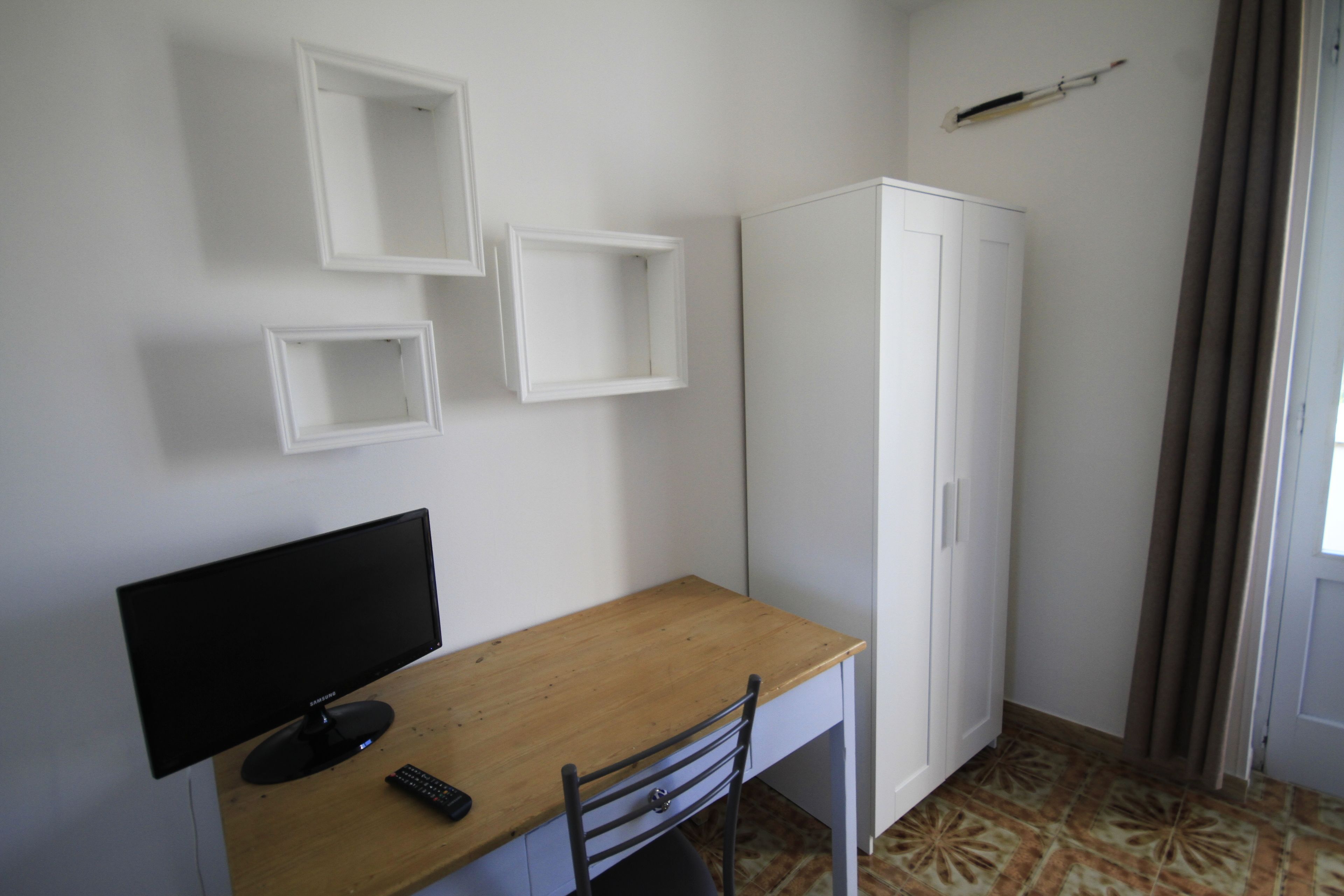 A room with a wooden desk, a black monitor, a white wardrobe, and a remote control.