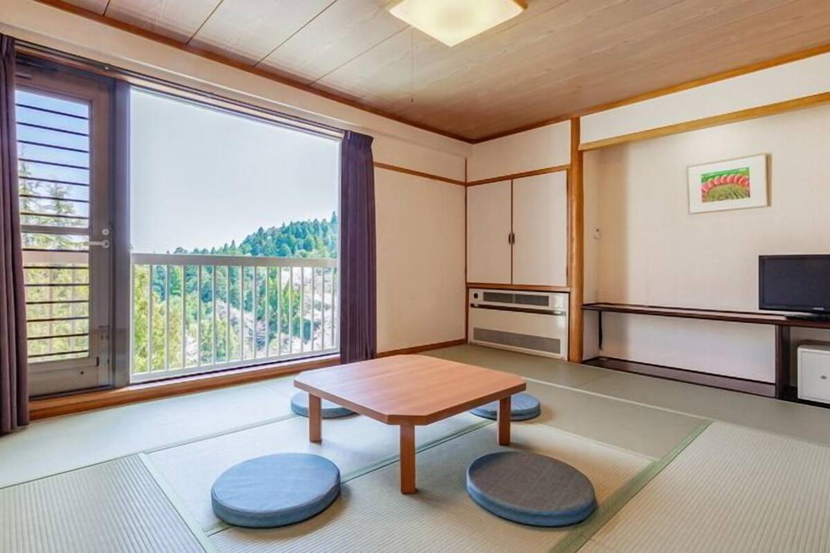 A room with tatami flooring, a low wooden table, tatami mats, a television, and a view of a forest through a sliding door.