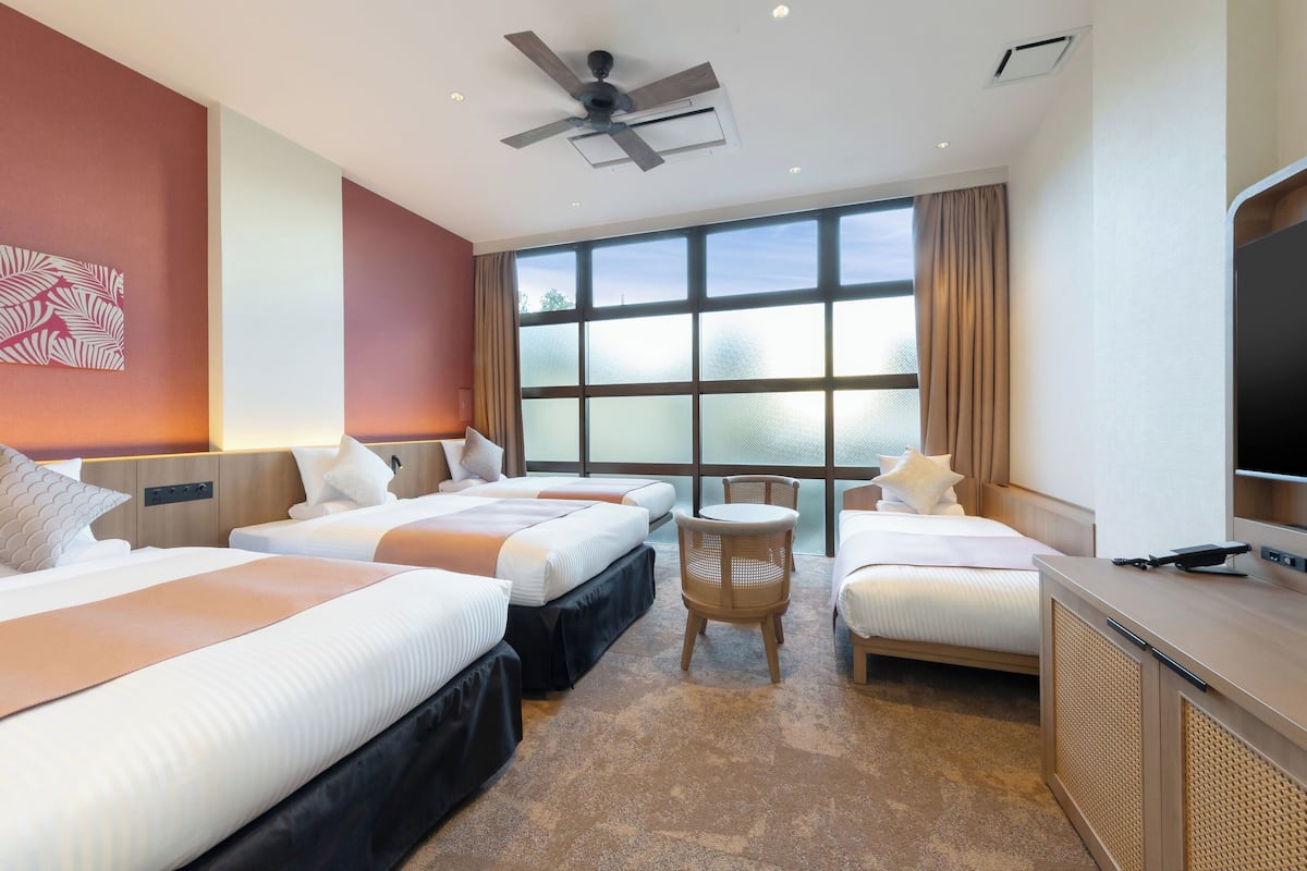 A hotel room with two beds, a television, a ceiling fan, and a large window with curtains.