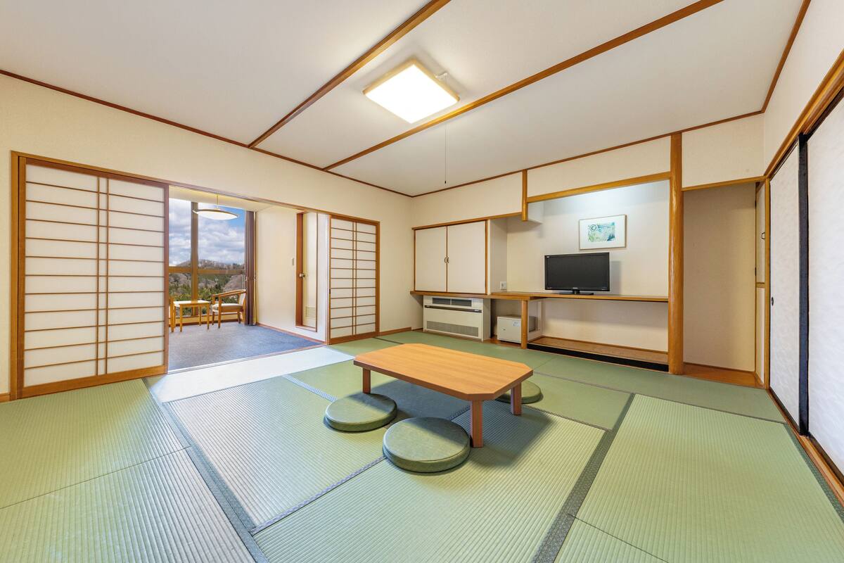 A traditional Japanese room with tatami flooring, a low wooden table, tatami mats, sliding doors, a television, and a view of the outdoors.
