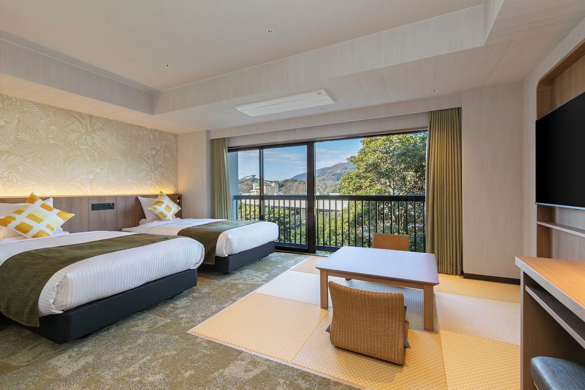 A hotel room with two beds, a dining table, a TV, and a balcony with a view.