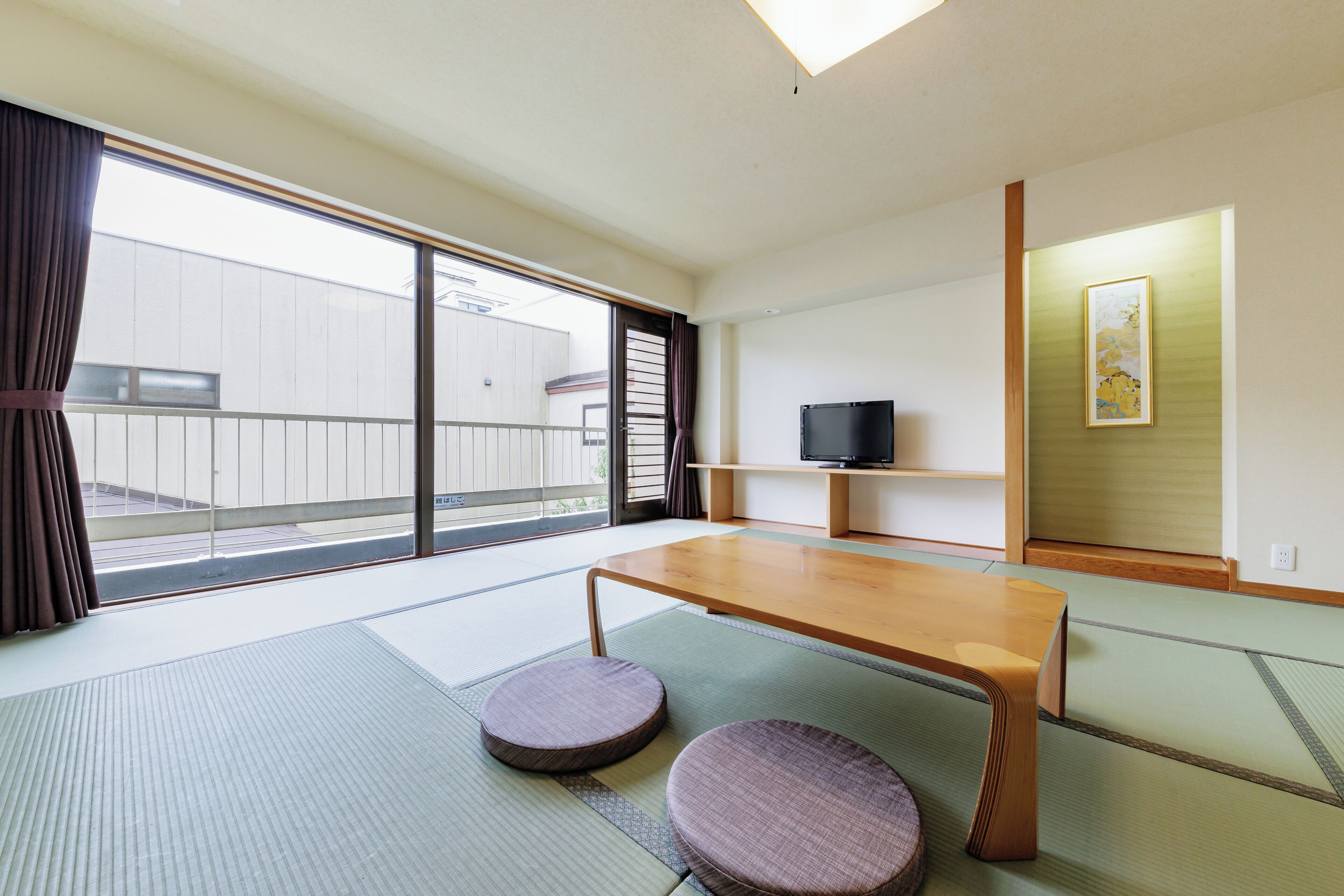 A room with tatami mats, a wooden table, two tatami cushions, a television on a shelf, and a sliding door to a balcony.