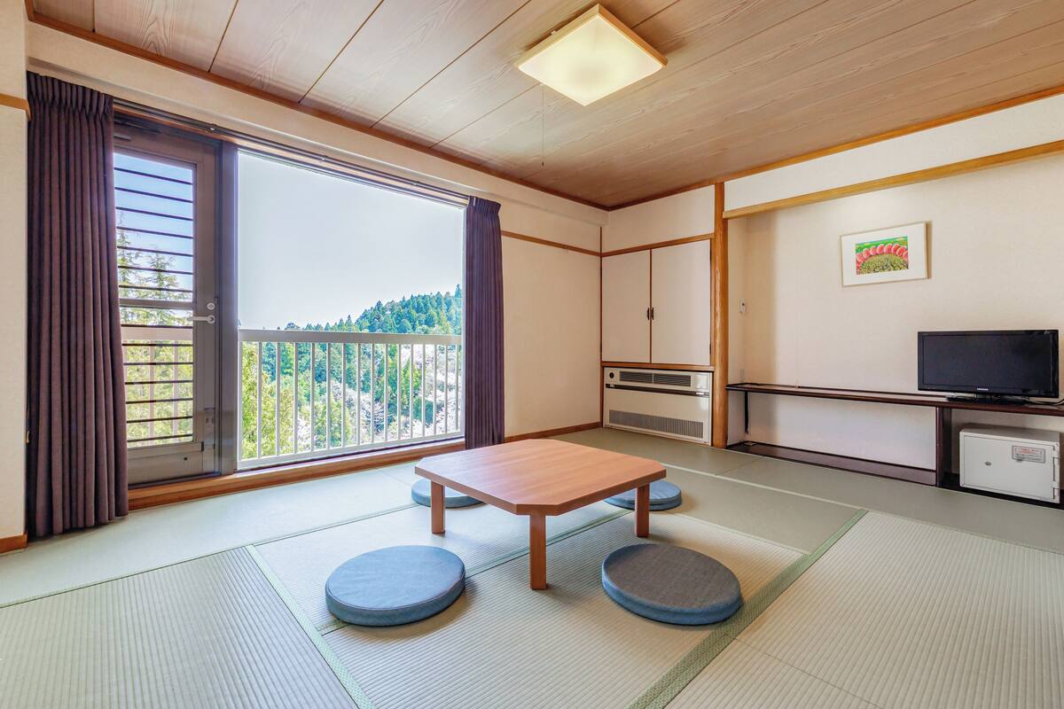 A traditional Japanese room with tatami flooring, a wooden table, cushions, a sliding door with a view, a TV, and a small cabinet.