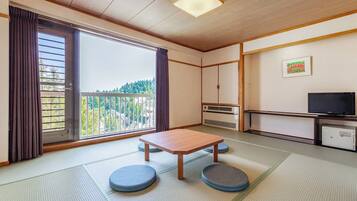 A traditional Japanese room with tatami flooring, a wooden table, cushions, a sliding door with a view, a TV, and a small cabinet.