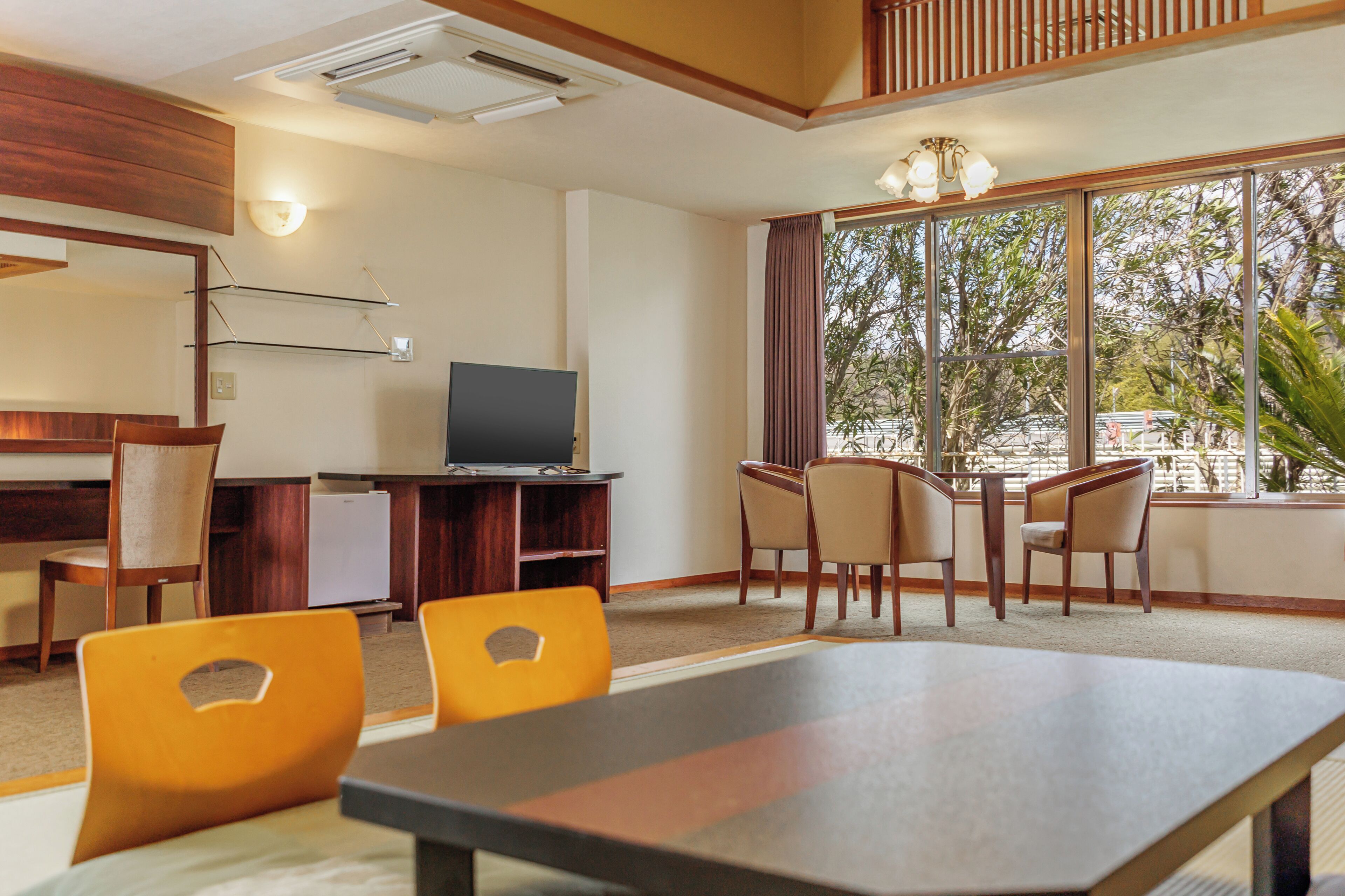 A hotel room with a dining area, a TV, and large windows with a view of trees.