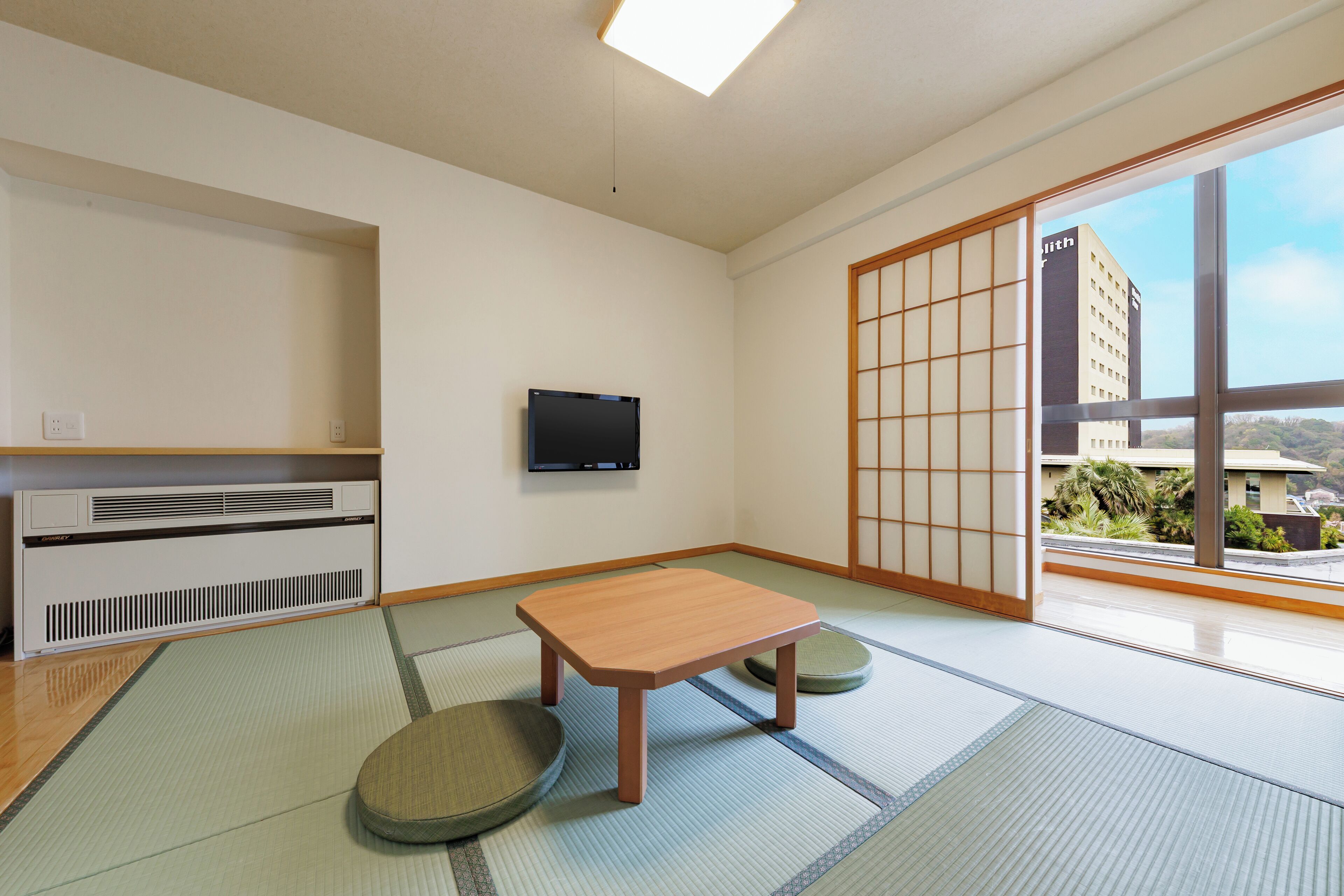A traditional Japanese room with tatami flooring, a low wooden table, a television, and a sliding door leading to a balcony with a view of a building and greenery.