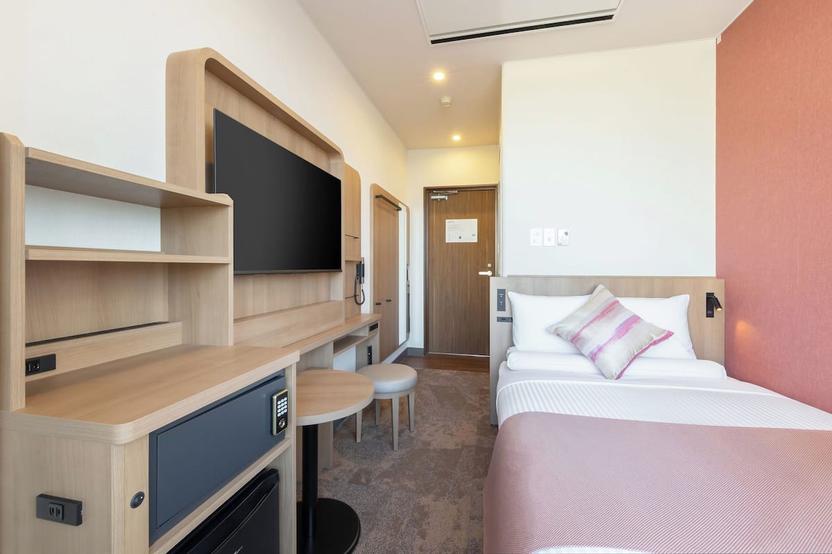 A hotel room with a bed, a desk with a flat-screen TV, a small table, and a chair.