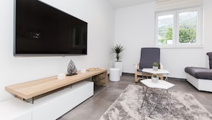 Een moderne woonkamer met een grote flatscreen-tv aan de muur, een houten salontafel, een grijze bank en een kleine bijzettafel met een plant.