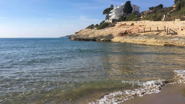 Una playa con agua cristalina, una orilla arenosa y un edificio blanco de varios pisos sobre una colina.