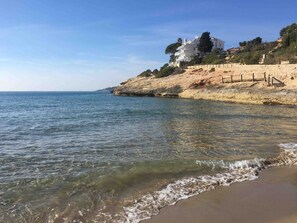 A beach with clear water, a sandy shore, and a white building with multiple floors on a hillside.