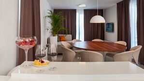 Habitación de hotel moderna con comedor que presenta una mesa redonda de madera, sillas color beige y una mesa blanca. Hay una planta en maceta, un sofá con un cojín naranja y un cuadro en la pared.
