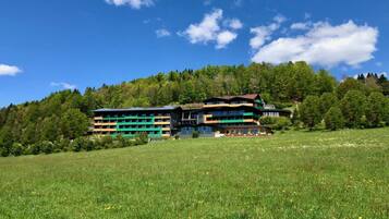 Un bâtiment moderne à plusieurs étages, entouré d’un champ verdoyant et d’une colline boisée.