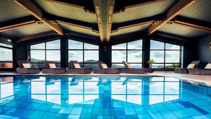 A large indoor swimming pool with clear blue tiles, surrounded by lounge chairs and large windows offering a view of the landscape. - Natura Hotel Bodenmais (Bodenmais)