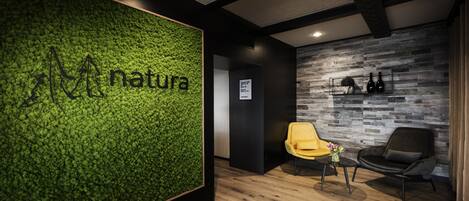 A reception area with a green wall displaying the word "natura," wooden flooring, a yellow chair, a black chair, and a small table with a vase.