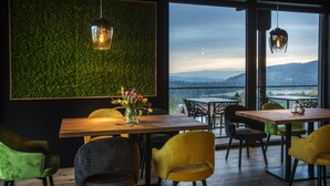 A dining area with a wooden table and chairs, a green wall with a textured pattern, and a view of mountains through large windows. - Natura Hotel Bodenmais (Bodenmais)