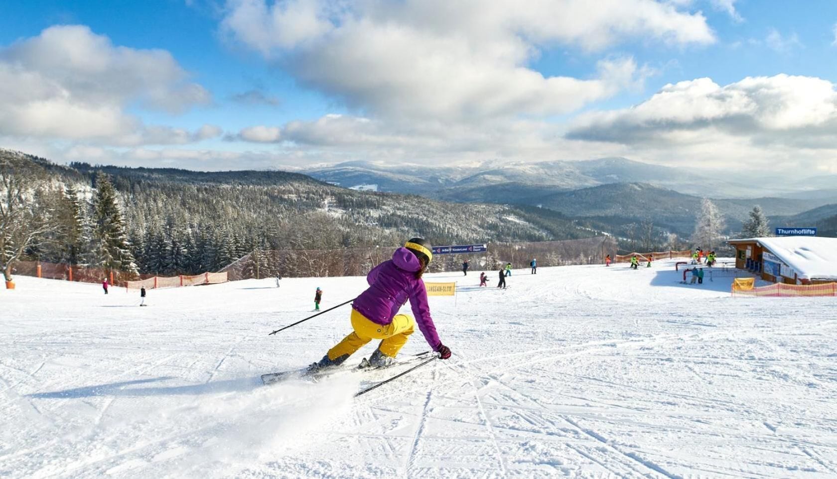 Une piste de ski avec des skieurs, un chalet en bois et, au loin, des montagnes enneigées.