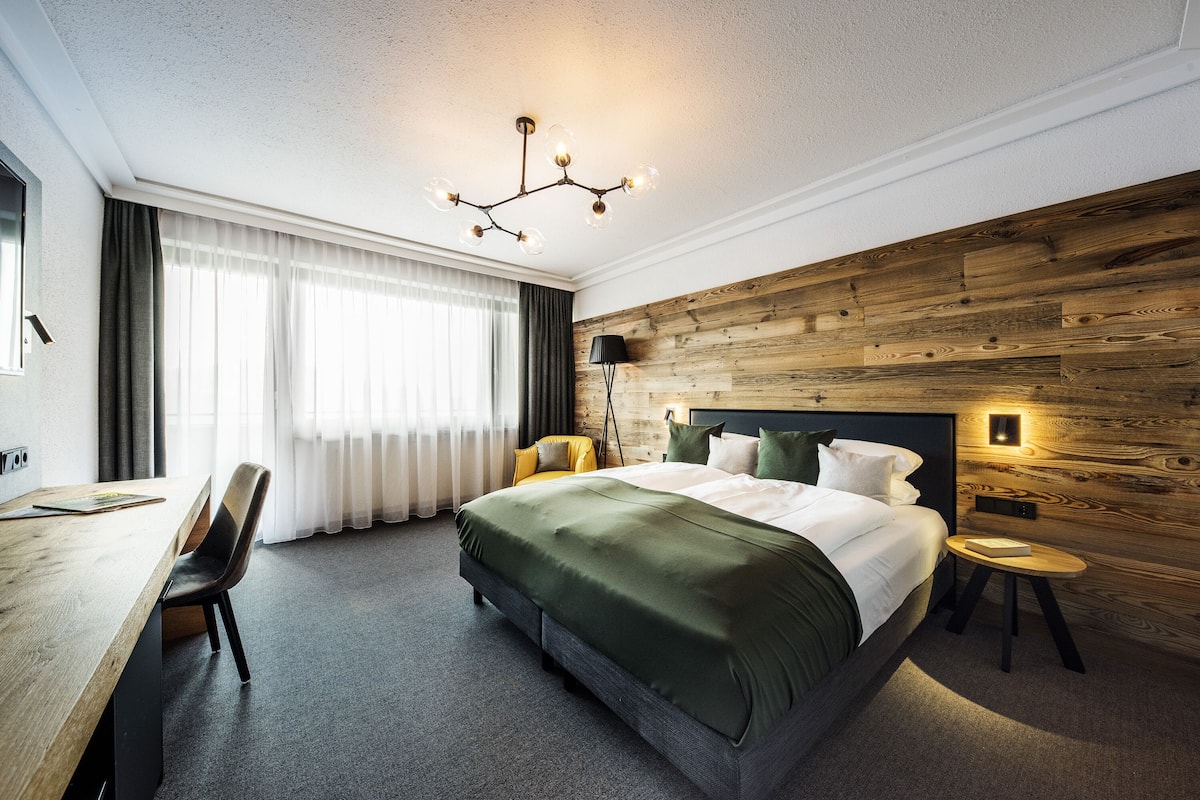 A modern hotel room with a large bed, a wooden accent wall, a desk with a chair, and a large window with curtains.