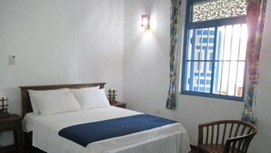Un dormitorio con cama, una silla, una mesita de noche, una lámpara de pared y una ventana con barrotes.