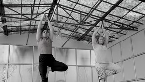 Dos personas practicando yoga en una habitación con un muro de ventanas con diseño de cuadrícula y estructura de techo metálica.