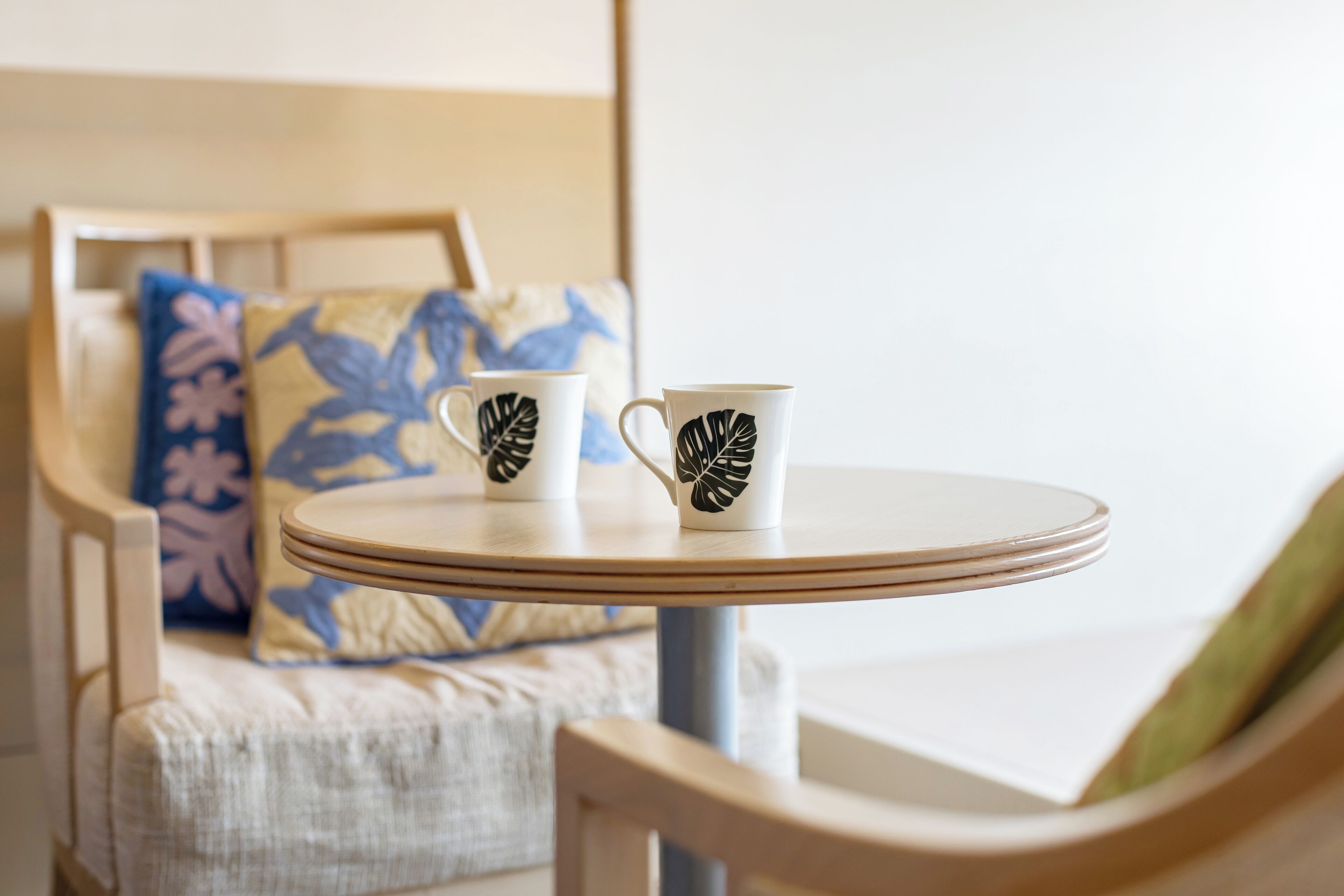 A round table with two white cups featuring black leaf designs, a wooden chair with a green cushion, and a patterned pillow in the background.
