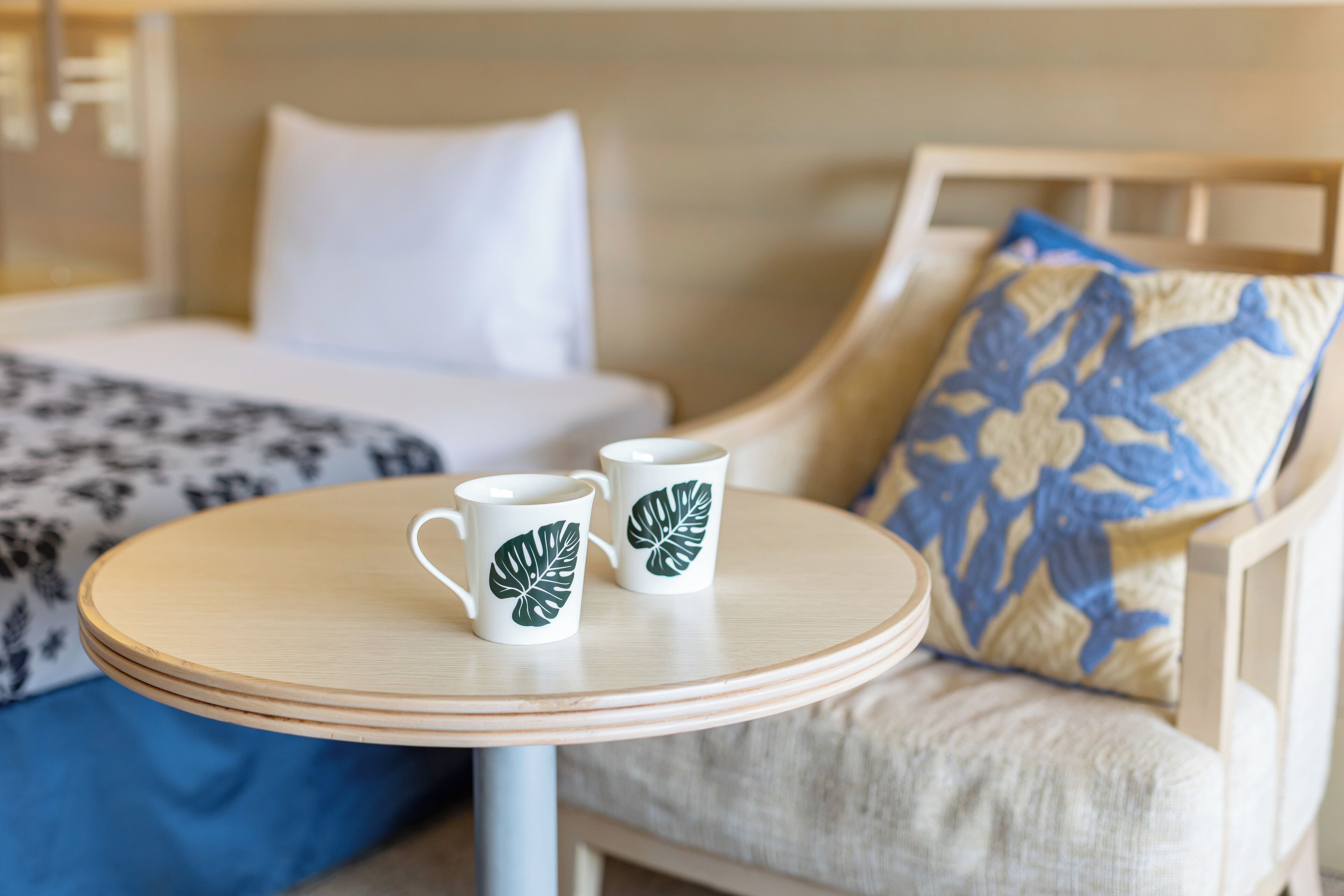 A round wooden table with two white mugs featuring green leaf designs, a beige sofa with a blue and beige patterned pillow, and a bed with white bedding and a black and white patterned blanket.