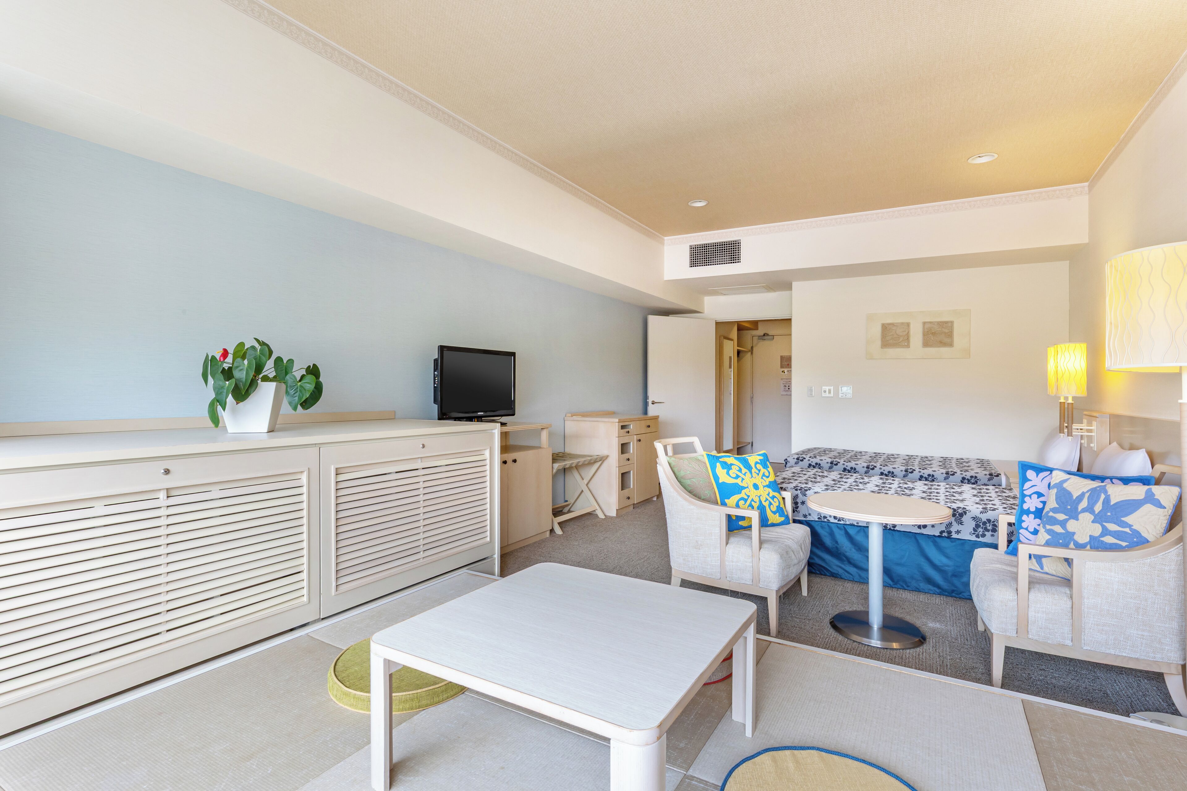A modern hotel room with a sofa, armchairs, a small table, a TV, and a plant.