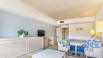 A modern hotel room with a sofa, armchairs, a small table, a TV, and a plant.