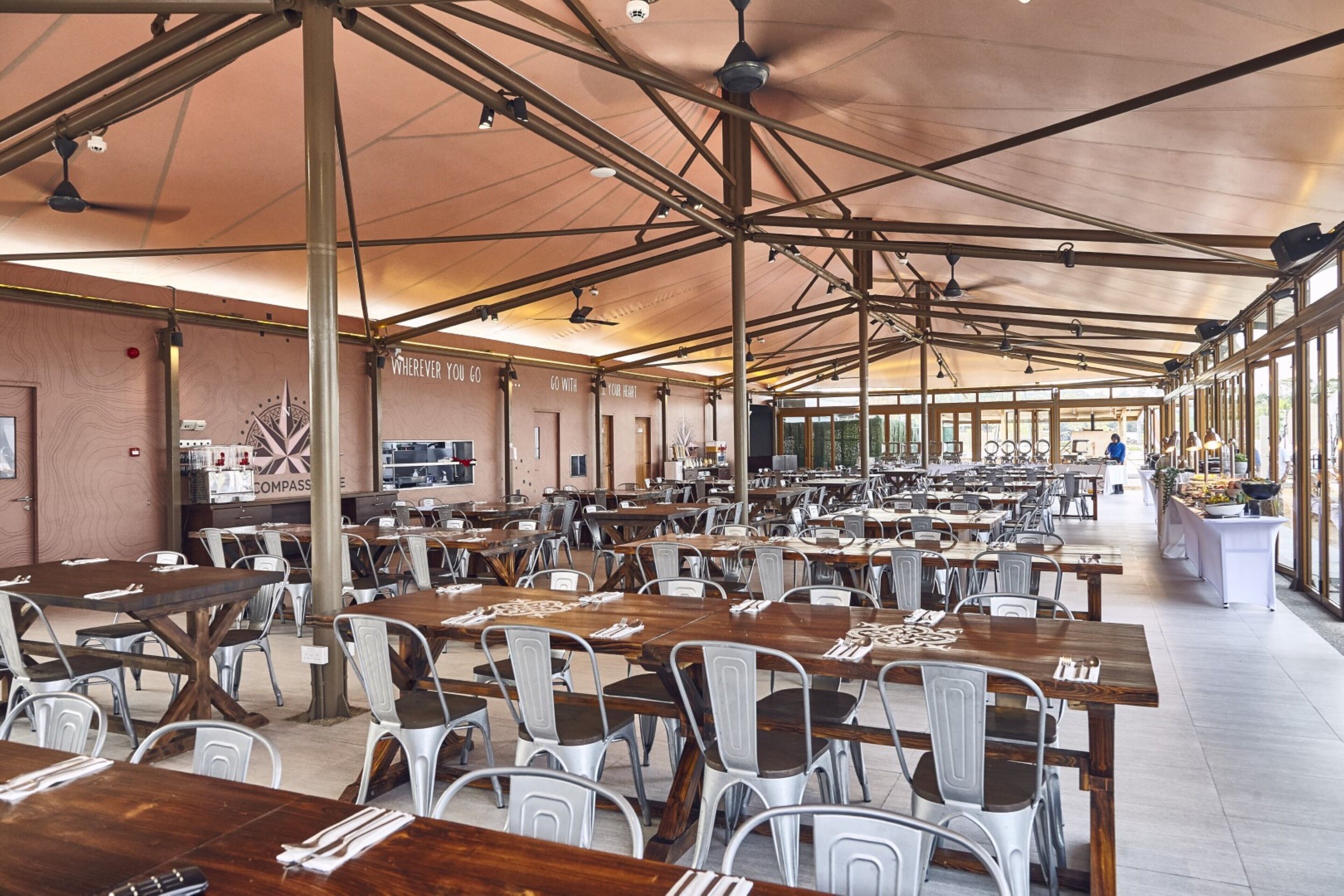 A spacious indoor dining area with multiple tables and chairs, high ceilings with exposed beams, and large windows allowing natural light.