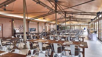 A spacious indoor dining area with multiple tables and chairs, high ceilings with exposed beams, and large windows allowing natural light.
