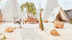 A series of white tents with conical roofs, arranged around a central wooden table and chairs, set on a paved area with rocks and cacti, under a large canopy structure. - The ANMON Resort Bintan- Glamping (Bintan)